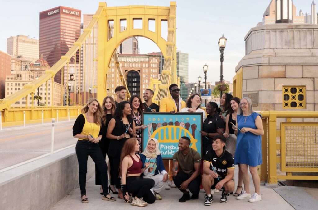 A group of people who painted a collection piece posing on a Pittsburgh Bridge with the artwork centered.