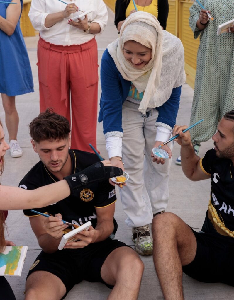Badawi holding palettes of paint for two people in soccer jerseys to paint with.