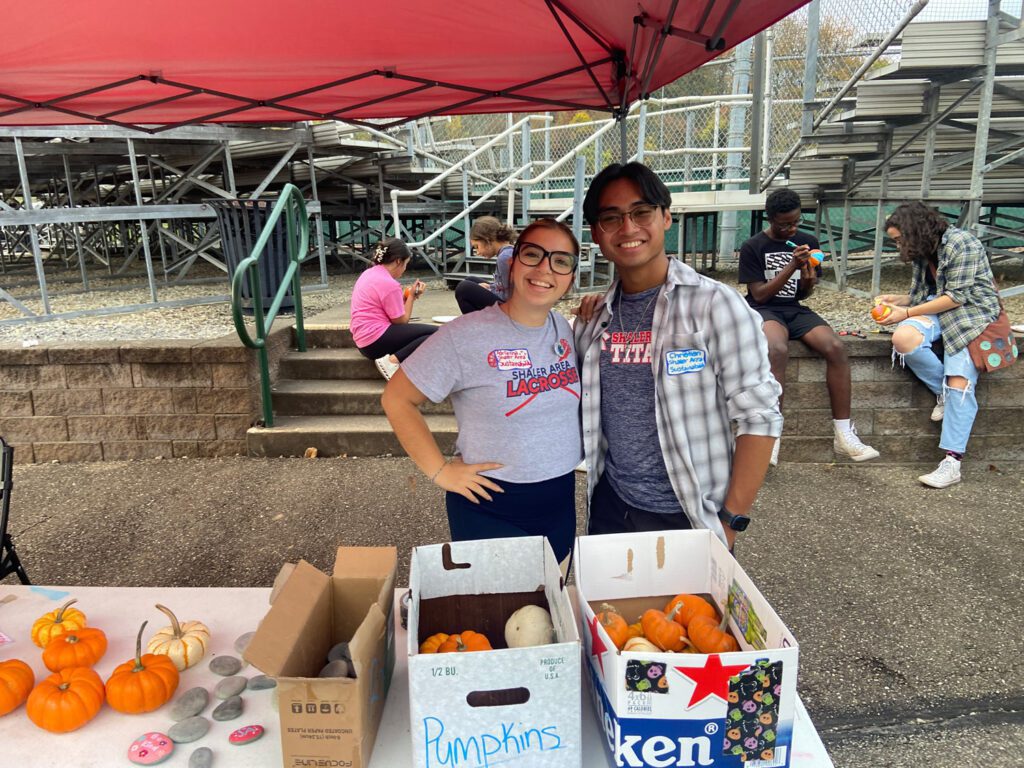 Two students under a tent in front of a table giving away pumpkins for decorating at a community event,