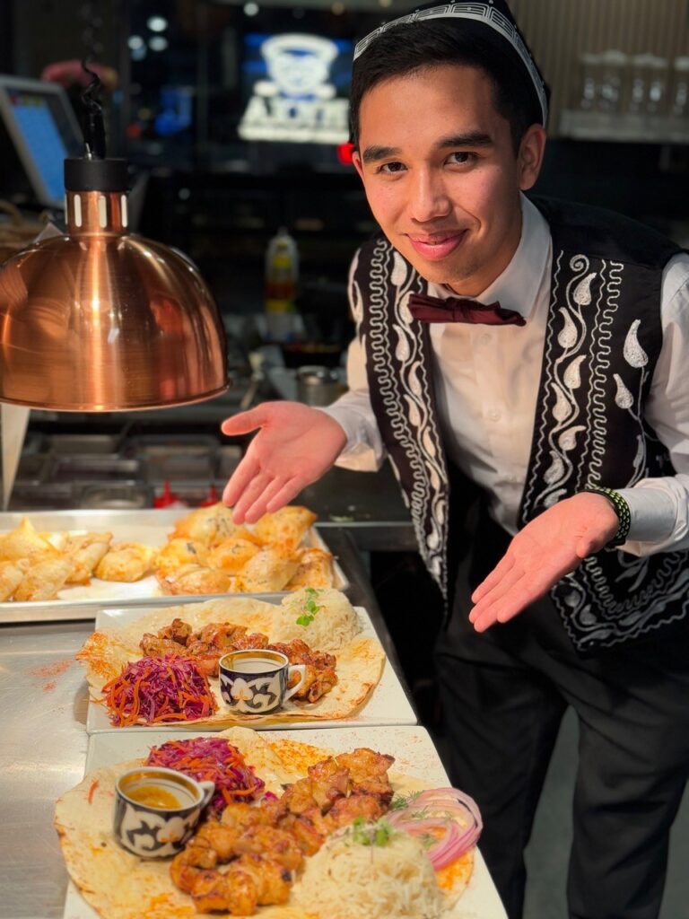 A server pointing to a display of meat, rice, and bread dishes under a heat lamp.