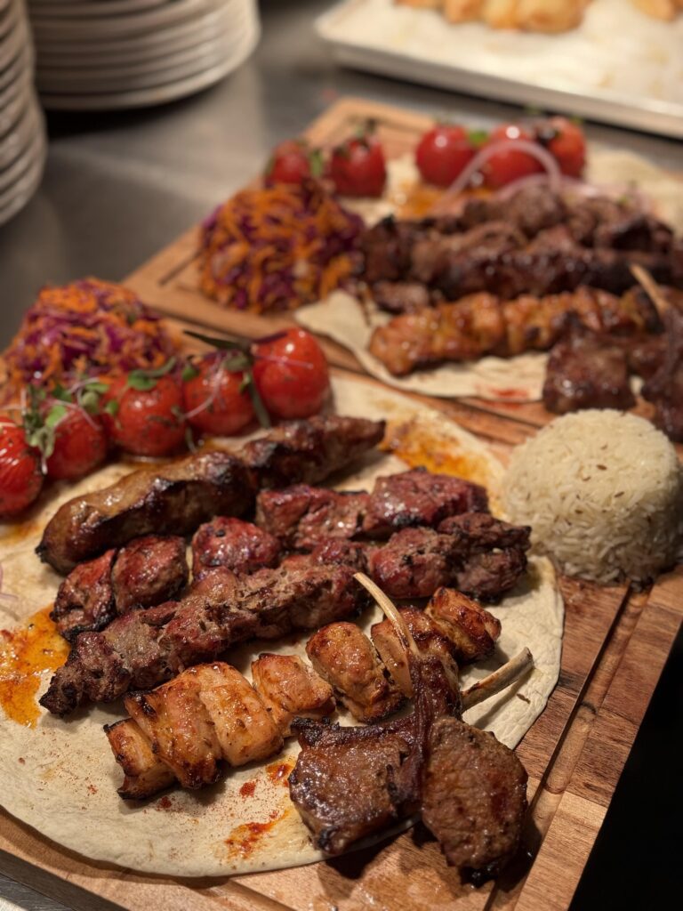 Kabobs in focus on pita, with rice and tomatoes on the vine, it is served on a wood platter.